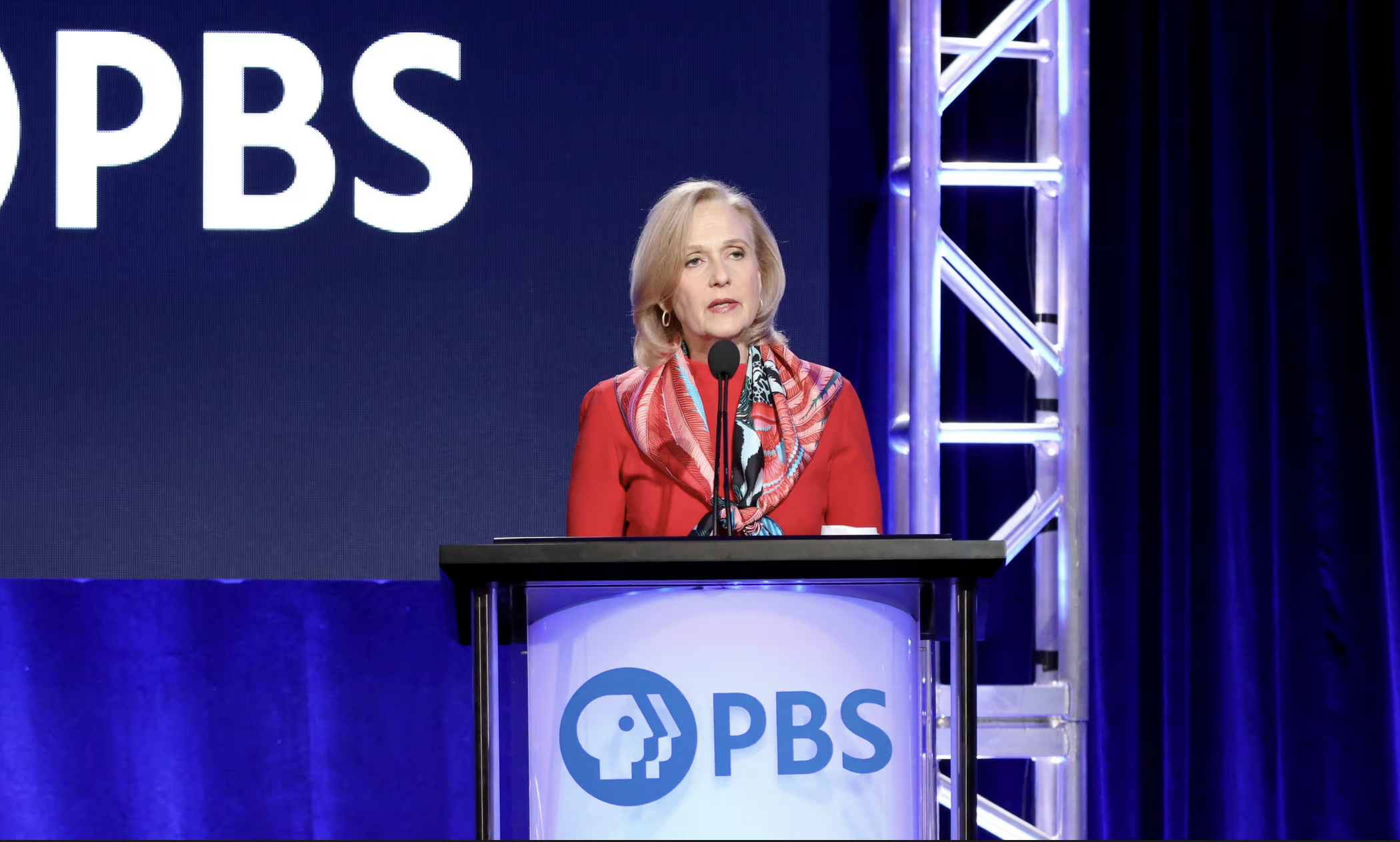 Person wearing red standing and speaking from behind a podium that says PBS.